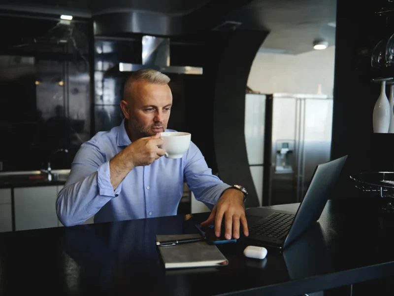 Man drinking coffee while processing payroll on a laptop.